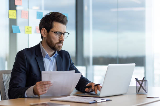 Serious and focused financier accountant on paper work inside office, mature man using calculator and laptop for calculating reports and summarizing accounts, businessman at work in suit