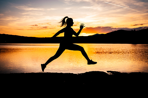 Silhouette of a sporty woman athlete jumping while exercising by the lake at sunset