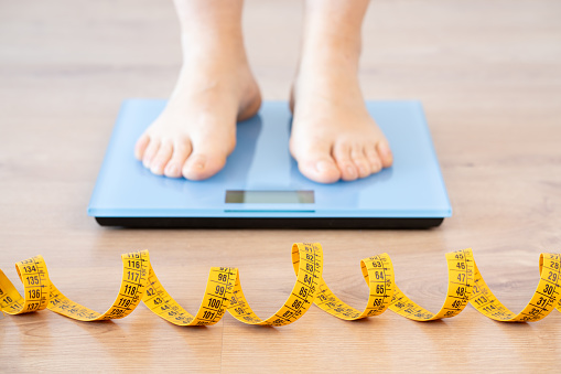Tape measure and woman feet standing on weigh scales.Diet, weight loss and feet of a woman on a scale for body check, measure and balance to lose weight on the floor. Obesity, scales and girl with tape