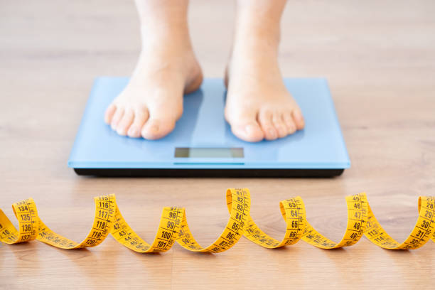 woman measuring her weight on the scale Tape measure and woman feet standing on weigh scales.Diet, weight loss and feet of a woman on a scale for body check, measure and balance to lose weight on the floor. Obesity, scales and girl with tape low quality of life stock pictures, royalty-free photos & images
