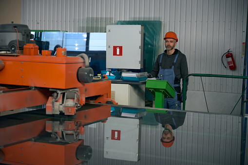 Specialist uses automatic table for cutting glass in a window production, a worker in a hard hat and protective gloves