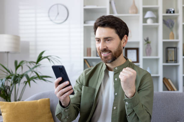 istock Mature bearded man joyously celebrating success while using smartphone on sofa at home 2035866894