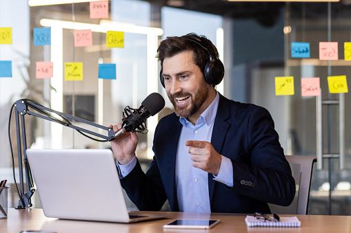 A businessman uses a professional microphone inside the office, to record a business training course podcast, a man in a business suit and headphones is happy and smiling at the laptop camera