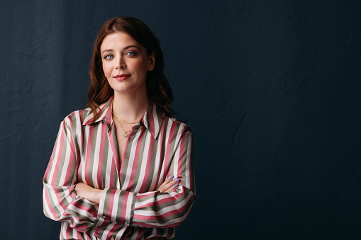 Portrait of a confident, professional woman with a slight smile, standing with her arms crossed, wearing a striped blouse against a dark background.