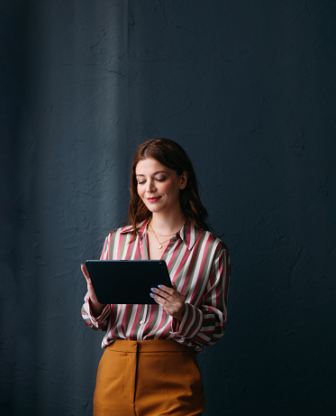Confident young female professional engaged in work on her tablet, wearing a striped shirt and smiling subtly.