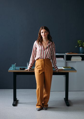 Professional young female in stylish outfit standing confidently by a modern workspace with a serene expression.