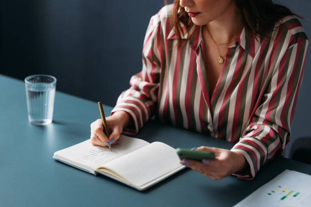 istock Focused Woman Writing in Notebook with Smartphone in Hand at Desk 2035915635