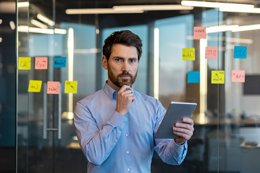 A mature businessman contemplates next moves while holding a tablet, standing in a modern office with sticky notes.