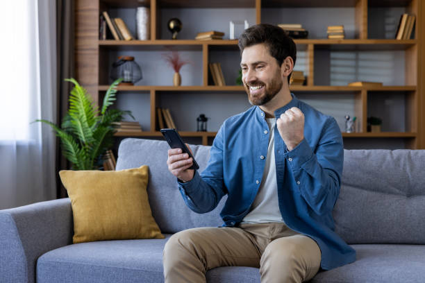 istock Smiling man enjoying success while using smartphone on couch at home 2035955432