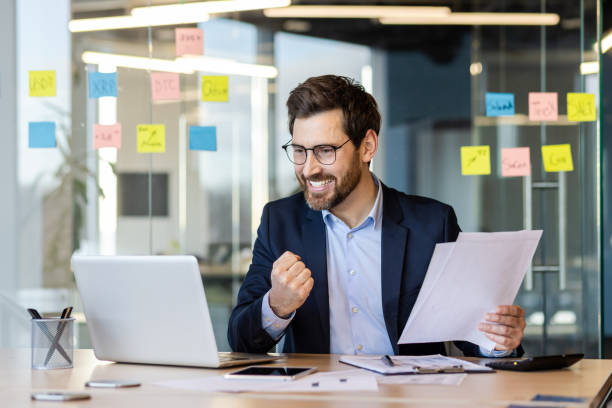 istock Successful accountant businessman in suit with documents, papers, reports in hands works inside office at workplace, male financier satisfied with results achievement celebrates victory uses laptop 2035986787