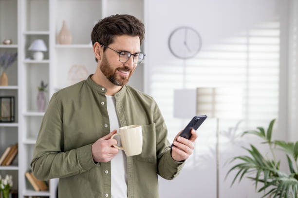 istock Smiling bearded man enjoying coffee while browsing smartphone in a bright modern home 2035999198