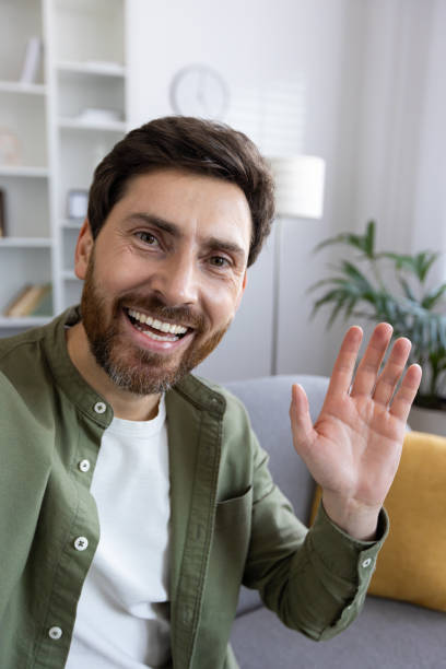 istock Cheerful bearded man waving hello, sitting comfortably at home on a sofa 2036009015