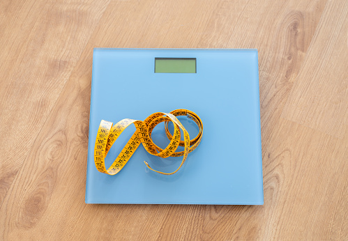 Closeup of a home scale and measuring tape on top of the blue scale on a wooden floor.