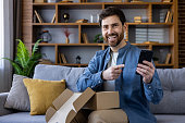 Smiling man with smartphone and open cardboard box enjoying online purchase at home