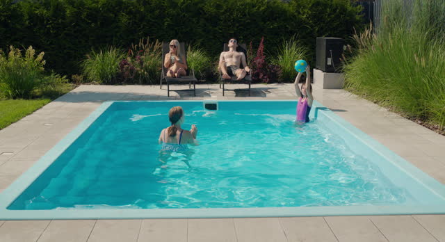 The family relaxes by their pool in the backyard of the house. Adults sunbathe on sun loungers, two children swim in the pool