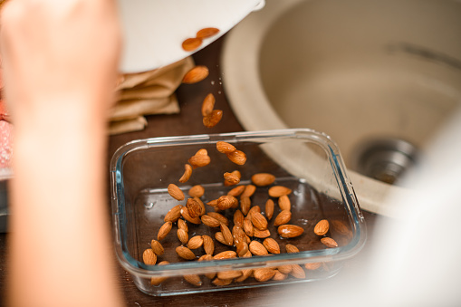 Female manicured baker's hands pouring almonds from a white deep bowl into a rectangular glass baking dish