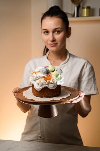 Baker in a white uniform holds a beautifully decorated Easter cake with an openwork round napkin on a stand