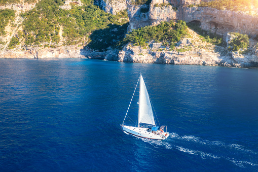 Aerial view of sailboat on blue sea at sunrise in summer. Travel in Sardinia, Italy. Tropical landscape with sailing boat, sea bay, rocky coast, green trees, water at dawn. Top view of yacht. Yachting