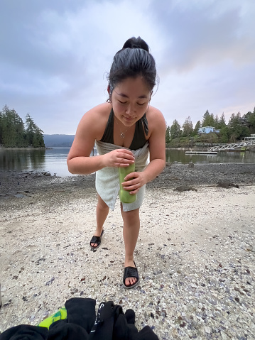 Multiracial young woman at a local neighborhood public beach drinking hot beverage in a reusable thermos while drying off at after a winter swim in the ocean.  North Vancouver, British Columbia, Canada.