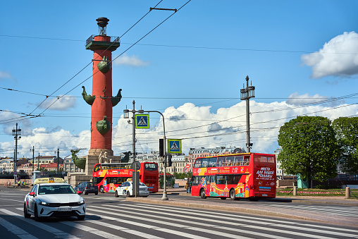Saint Petersburg. Russia - June 02, 2023: Rostral column (lighthouse) on embankment Neva river. Vasilyevskiy island in Saint Petersburg, blue cloudy sky, bus and car on the road.\n.