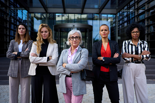 Group of empowered diverse race and ages business women entrepreneurs standing in formal wear with arms crossed looking at camera. Serious and successful adult female professionals co-workers outdoors