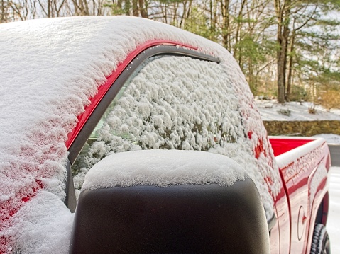 Coating of snow over a red truck windshield, window, and side view mirror. Heavy wet snow covers a red vehicle in New England snow country. A winter theme with copy space.