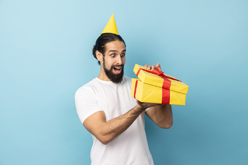 Portrait of man with beard wearing white T-shirt and party cone unpacking present, looking inside box with shocked and scared expression, birthday gift. Indoor studio shot isolated on blue background.