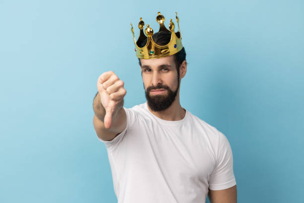 istock Man in gold crown standing looking at camera with disappointed expression, showing thumb down. 2036397761
