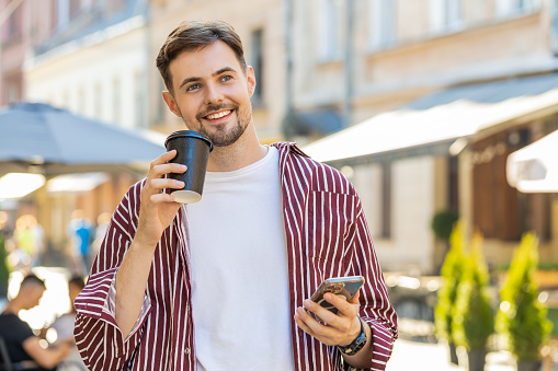 Happy brunette bearded man enjoying morning coffee hot drink and smiling outdoors. Relaxing taking a break. Guy walking in urban city sunshine street, drinking coffee to go. Town lifestyles outside