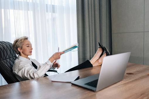 Relaxed middle-aged female CEO putting feet on desk with laptop. Confident successful business woman thinking about career growth relaxing in cabinet. Concept relaxation at workplace