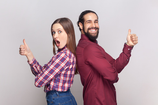 Portrait of young adult woman and man standing back to back together showing thumbs up, like gesture, positive feedback, looking at camera. Indoor studio shot isolated on gray background.