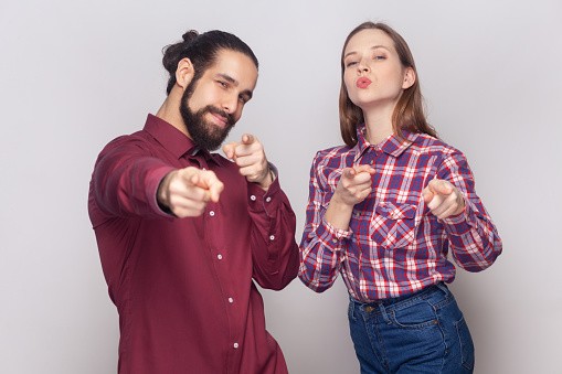 Portrait of satisfied delighted cute woman and man standing together pointing at camera, selecting you, expressing positive emotions. Indoor studio shot isolated on gray background.