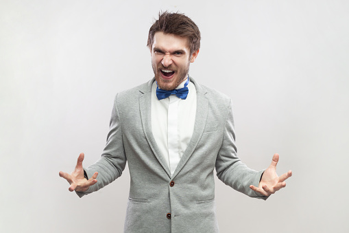 Portrait of angry aggressive handsome bearded man standing with raised arms, screaming, expressing hate and anger, wearing grey suit and blue bow tie. Indoor studio shot isolated on gray background.