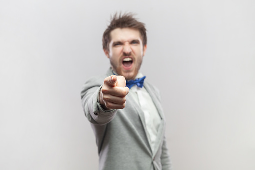 Portrait of angry aggressive bearded man standing pointing at you, choosing you, arguing and screaming, wearing grey suit and blue bow tie. Indoor studio shot isolated on gray background.