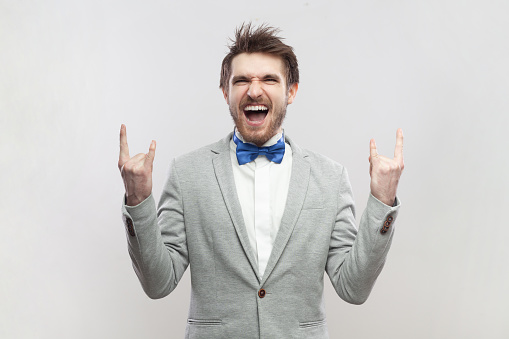 Portrait of crazy funny excited handsome bearded man standing with rock and roll gesture, screaming with excitement, wearing grey suit and blue bow tie. Indoor studio shot isolated on gray background.