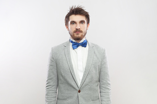 Portrait of serious attentive concentrated bearded man standing looking at camera with strict bossy expression, wearing grey suit and blue bow tie. Indoor studio shot isolated on gray background.