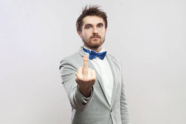 Rude impolite handsome bearded man standing showing middle finger, arguing with somebody. Portrait of rude impolite handsome bearded man standing showing middle finger, arguing with somebody, wearing grey suit and blue bow tie. Indoor studio shot isolated on gray background. asshole stock pictures, royalty-free photos & images