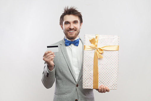 Portrait of joyful cheerful smiling bearded man standing with present box and showing credit card, wearing grey suit and blue bow tie. Indoor studio shot isolated on gray background.