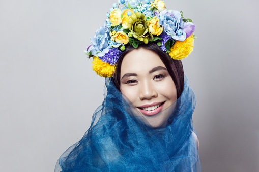 Closeup portrait of smiling happy sensual woman fashion model with beautiful makeup with floral hat and blue veil, looking at camera. Indoor studio shot isolated on gray background,