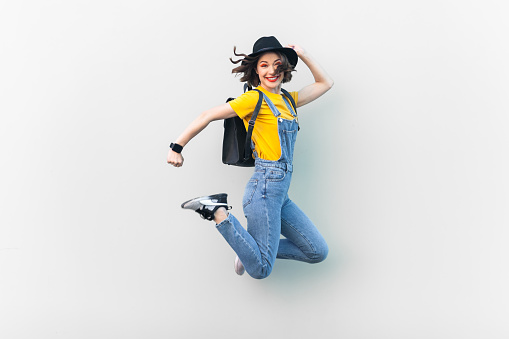 Portrait of attractive overjoyed hipster woman in blue denim overalls, yellow T-shirt and black hat, jumping, being in good mood, expressing happiness. Indoor studio shot isolated on gray background.