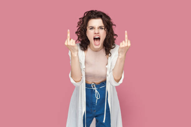 Rude impolite woman with curly hair standing showing middle fingers, screaming with hate and anger. Portrait of rude impolite woman with curly hair wearing casual style outfit standing showing middle fingers, screaming with hate and anger. Indoor studio shot isolated on pink background. asshole stock pictures, royalty-free photos & images