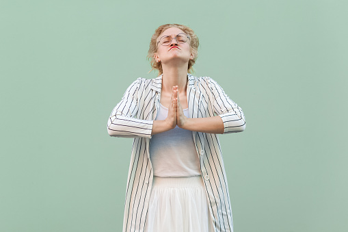 Portrait of attractive hopeful young adult blonde woman wearing striped shirt and skirt, keeps hands in praying gesture, asking to forgive her. Indoor studio shot isolated on light green background.