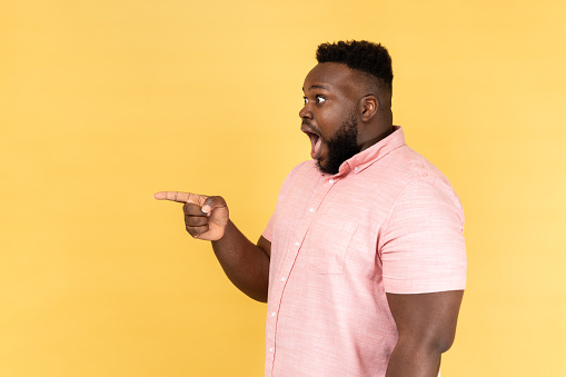 Side view portrait of shocked amazed man wearing pink shirt standing pointing away at copy space for advertisement or promotion. Indoor studio shot isolated on yellow background.
