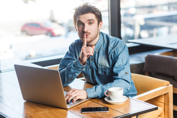 Man freelancer working on laptop, showing shh gesture, looking at camera, asking not to make noise