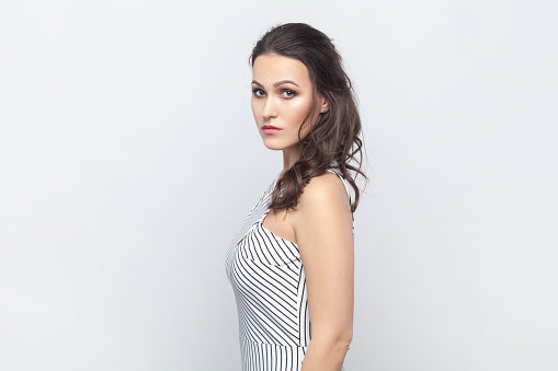 Side view portrait of strict bossy brunette woman standing looking at camera, having serious facial expression, wearing striped dress. Indoor studio shot isolated on gray background.