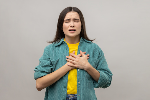 Portrait of sick woman suffering from pain in chest, grimacing with closed eyes, risk of heart attack or breast cancer, wearing casual style jacket. Indoor studio shot isolated on gray background.