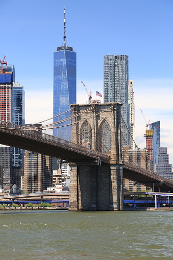 Manhattan bridge in New York. In the background, modern skyscraper One World Trade Center, construction cranes, flag of the United States of America. Sunny day