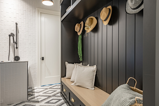 Interior photograph of a mudroom with a pet food station, dog shower beautiful tile floor and a shiplap walled coat rack with a bench seat and straw hats hanging on hooks. The bench has pillows and there is a stuffed Dalmatian dog sitting on the counter above the dog food bowls.
