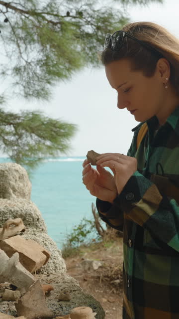Vertical video. Fragments of ancient clay pottery in the ruins of the city of Phaselis by the sea. A young woman studies them.