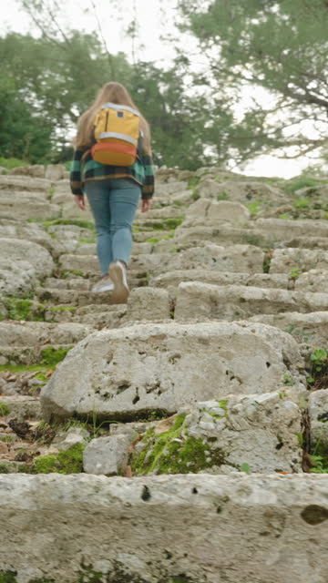Vertical video. A female tourist with a backpack climbs the steps of an ancient Greek amphitheater overgrown with forest for thousands of years.